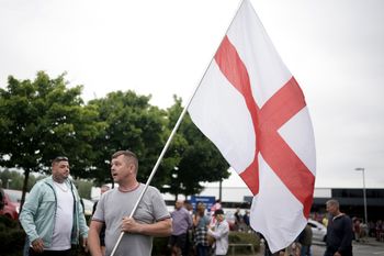ROTHERHAM, ENGLAND - AUGUST 4: An anti-immigration protester holds a St. George's cross flag outside of the Holiday Inn Express in Manvers, which is being used as an asylum hotel, on August 4, 2024 in Rotherham, United Kingdom. Yesterday saw widespread violence as Far-right agitators in Liverpool and Manchester rioted and looted shops. Police were attacked and injured and dozens of arrests were made. (Photo by Christopher Furlong/Getty Images)