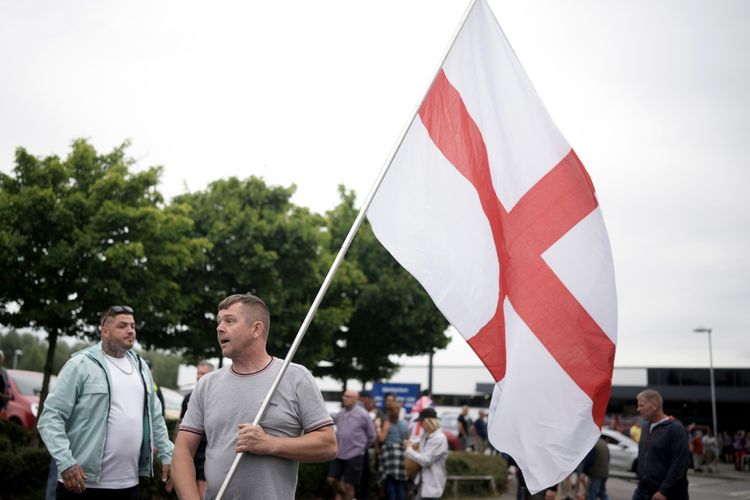 ROTHERHAM, ENGLAND - AUGUST 4: An anti-immigration protester holds a St. George's cross flag outside of the Holiday Inn Express in Manvers, which is being used as an asylum hotel, on August 4, 2024 in Rotherham, United Kingdom. Yesterday saw widespread violence as Far-right agitators in Liverpool and Manchester rioted and looted shops. Police were attacked and injured and dozens of arrests were made. (Photo by Christopher Furlong/Getty Images)