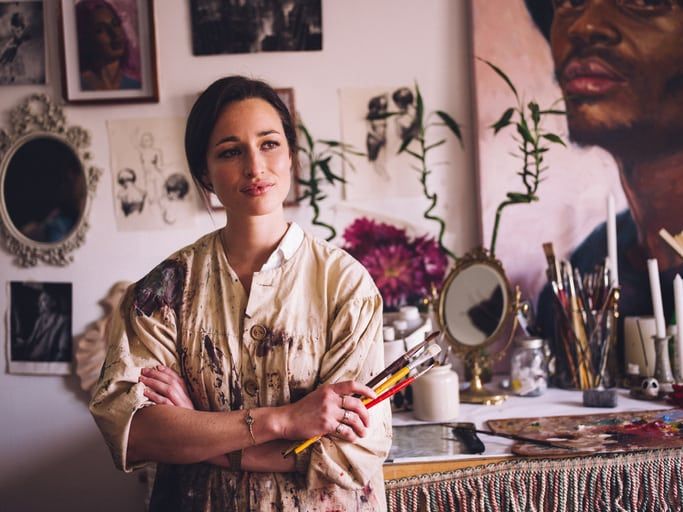 Beautiful young woman artist looking away thoughtfully with her arms crossed and holding some paintbrushes in her creative studio wearing an old-fashioned artist's smock which is covered with paint marks